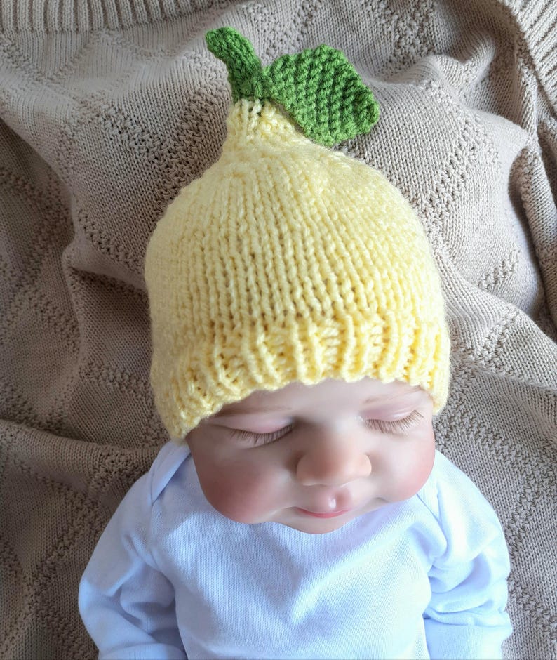 May include: A knitted baby hat in the shape of a lemon, with a green leaf detail. The hat is yellow and sits on a baby wearing a white onesie. The baby is lying on a beige blanket.