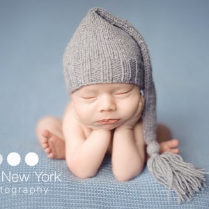 May include: A newborn baby wearing a gray knitted hat with a long tassel. The baby is lying on a blue blanket with its hands tucked under its chin.