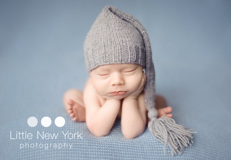 May include: A newborn baby wearing a gray knitted hat with a long tassel. The baby is lying on a blue blanket with its hands tucked under its chin.
