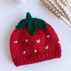 May include: A red knit strawberry hat with a green leaf crown and white seed accents. The hat is a novelty item, likely for a baby or child. The hat is made of a soft, textured yarn.