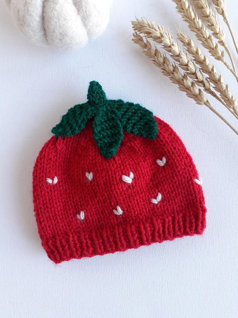 May include: A red knit strawberry hat with a green leaf crown and white seed accents. The hat is a novelty item, likely for a baby or child. The hat is made of a soft, textured yarn.