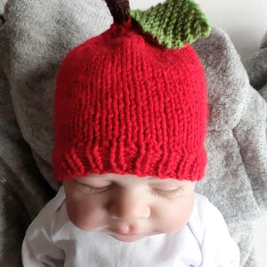 May include: A red knitted baby hat shaped like an apple, featuring a brown stem and a green leaf. The hat is worn by a baby wearing a white onesie. The hat is a unique and playful accessory.