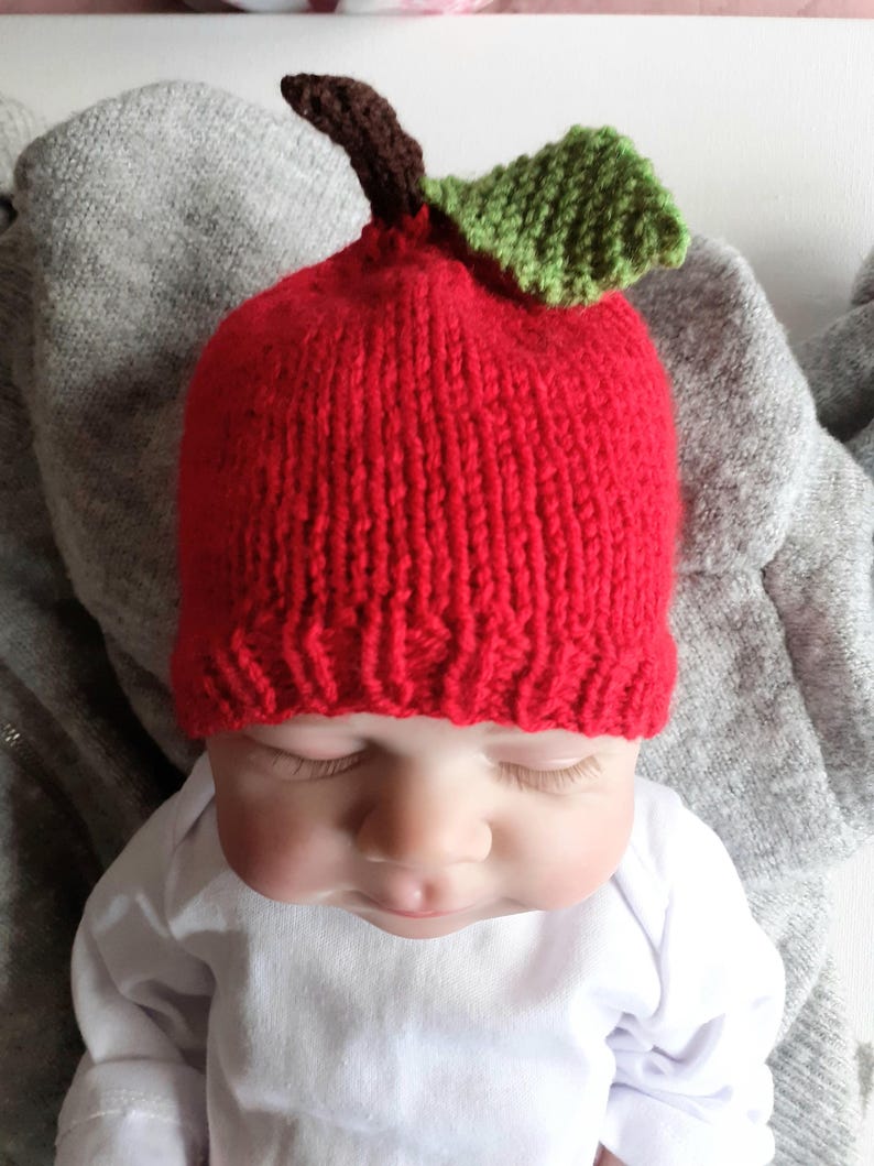 May include: A red knitted baby hat shaped like an apple, featuring a brown stem and a green leaf. The hat is worn by a baby wearing a white onesie. The hat is a unique and playful accessory.