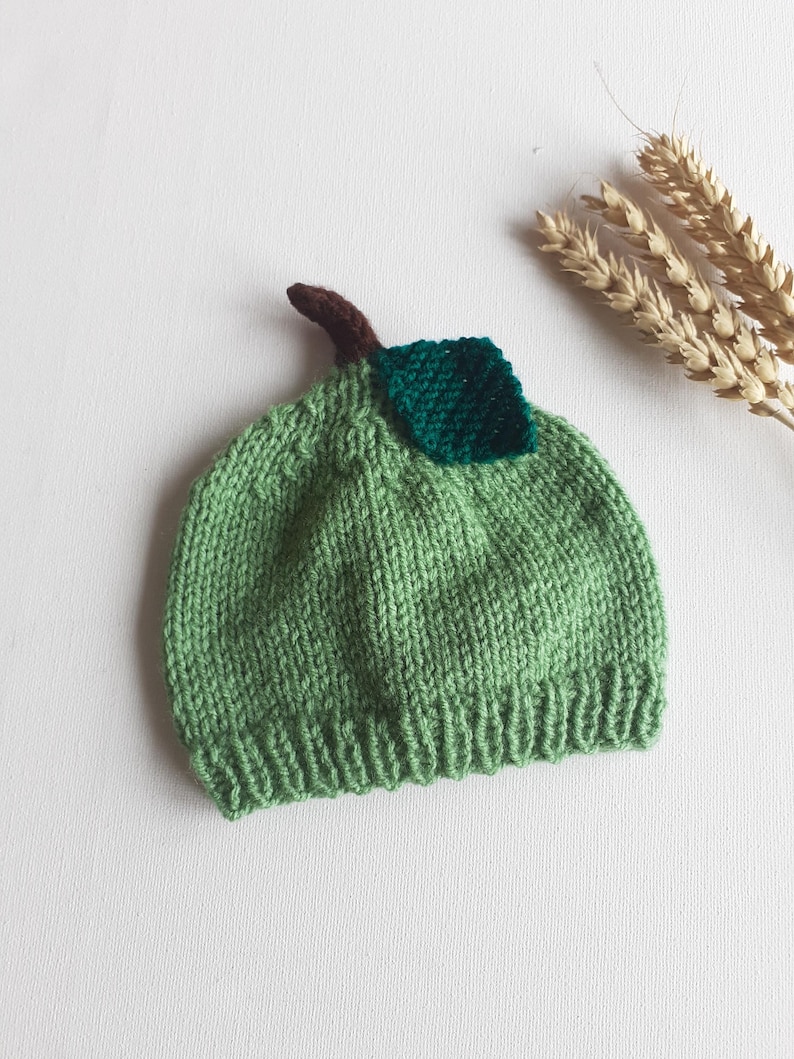 May include: A knitted green baby hat shaped like a pear, with a brown stem and a green leaf. The hat is made of soft yarn and is designed to fit a baby's head. The hat is a unique and whimsical accessory.