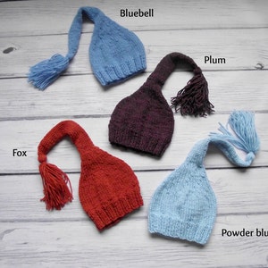 May include: Four knitted baby hats with long tails and tassels in the colors bluebell, plum, fox, and powder blue.