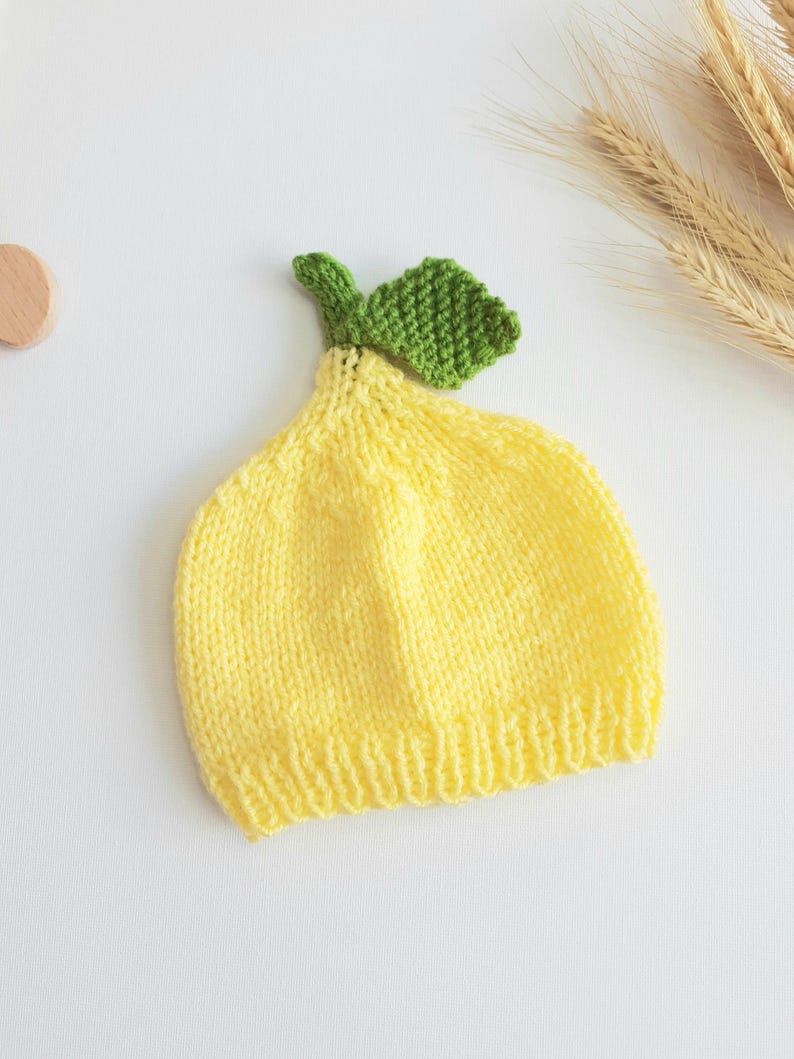 May include: A knitted baby hat in the shape of a lemon. The hat is primarily yellow with a green leaf and stem detail at the top. The hat is made of soft yarn and is designed to fit a baby's head.