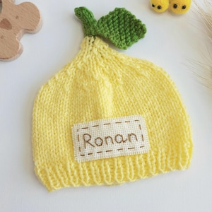 May include: A yellow knitted baby hat shaped like a lemon, with a green leaf and the name "Ronan" embroidered on a patch. The hat is next to two small yellow bee decorations.