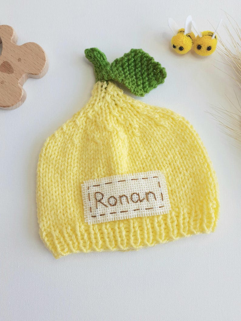 May include: A yellow knitted baby hat shaped like a lemon, with a green leaf and the name "Ronan" embroidered on a patch. The hat is next to two small yellow bee decorations.