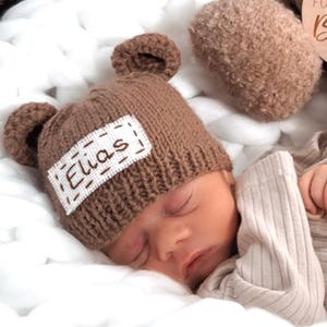 May include: A brown knitted baby hat with bear ears and a white patch that says "Elias" in brown embroidery.
