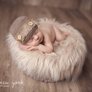 K&ouml;nnte beinhalten: Ein neugeborenes Baby tr&auml;gt eine braune Strickm&uuml;tze mit einem Leinenband und Holzkn&ouml;pfen und schl&auml;ft auf einem flauschigen wei&szlig;en Fellteppich.