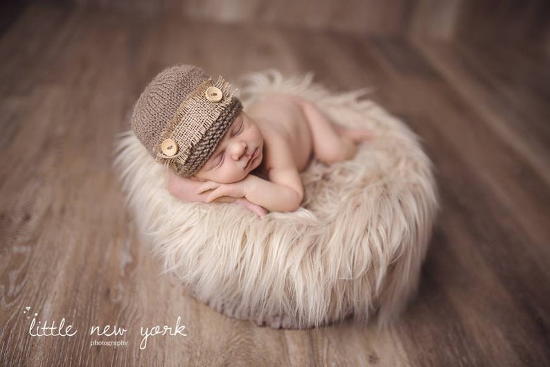 K&ouml;nnte beinhalten: Ein neugeborenes Baby tr&auml;gt eine braune Strickm&uuml;tze mit einem Leinenband und Holzkn&ouml;pfen und schl&auml;ft auf einem flauschigen wei&szlig;en Fellteppich.
