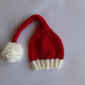 May include: A long, red knitted hat with a white ribbed brim and a long, drooping tail that ends in a large, fluffy white pom-pom. The hat features a classic stocking cap shape with a textured knit pattern throughout the red and white sections.