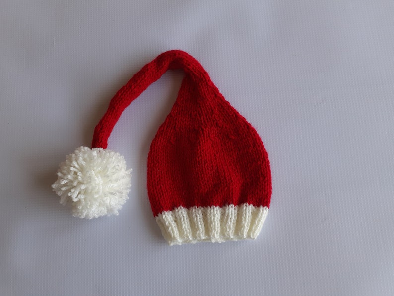 May include: A long, red knitted hat with a white ribbed brim and a long, drooping tail that ends in a large, fluffy white pom-pom. The hat features a classic stocking cap shape with a textured knit pattern throughout the red and white sections.