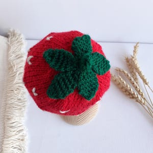 May include: A red knitted hat shaped like a strawberry, with a green leaf design on top and white seed details. The hat is designed to look like a strawberry. The hat is a fun accessory.