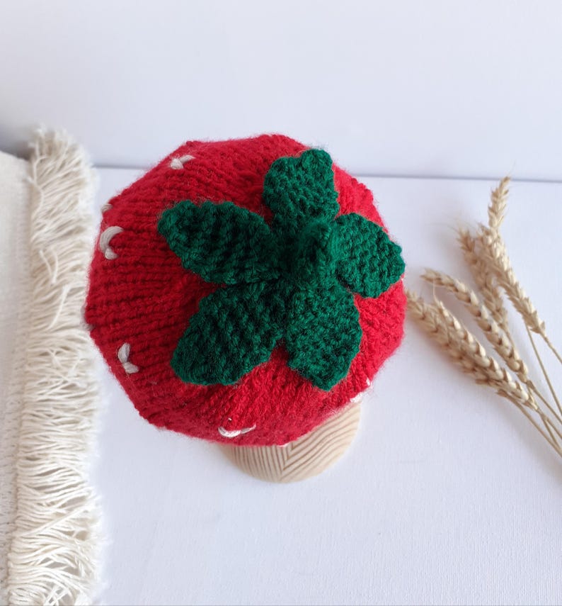 May include: A red knitted hat shaped like a strawberry, with a green leaf design on top and white seed details. The hat is designed to look like a strawberry. The hat is a fun accessory.