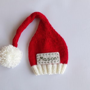 May include: A red knitted hat with a long, drooping tail and a white pom-pom at the end. The hat features a white ribbed brim and a rectangular fabric patch stitched onto the front with the name Mason embroidered in dark green thread.