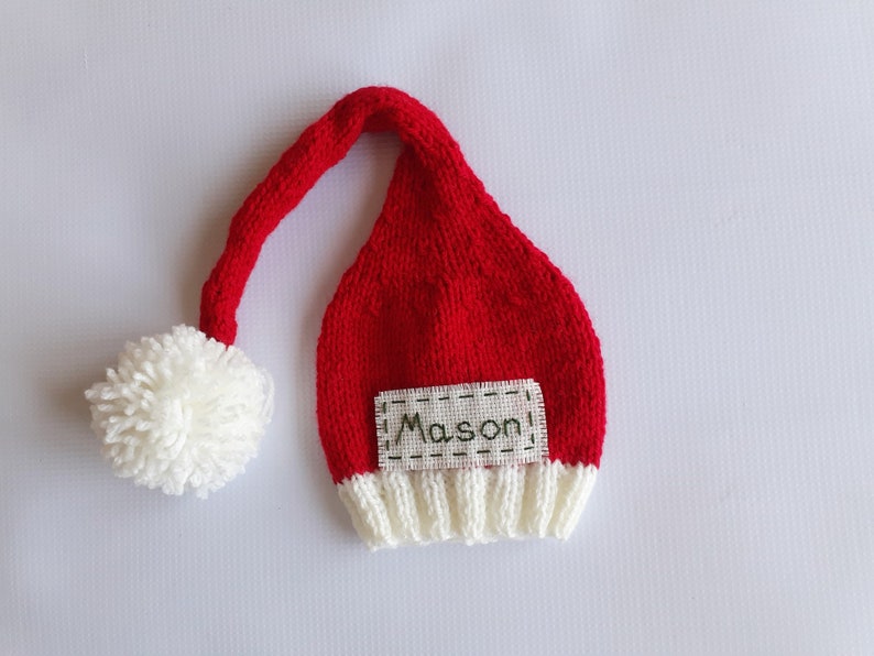 May include: A red knitted hat with a long, drooping tail and a white pom-pom at the end. The hat features a white ribbed brim and a rectangular fabric patch stitched onto the front with the name Mason embroidered in dark green thread.