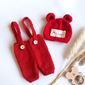 May include: A bright red knitted overall set with two light-colored buttons on the front straps and a matching red knitted hat featuring two small rounded ears and a white rectangular patch with the name Mason embroidered in red thread.
