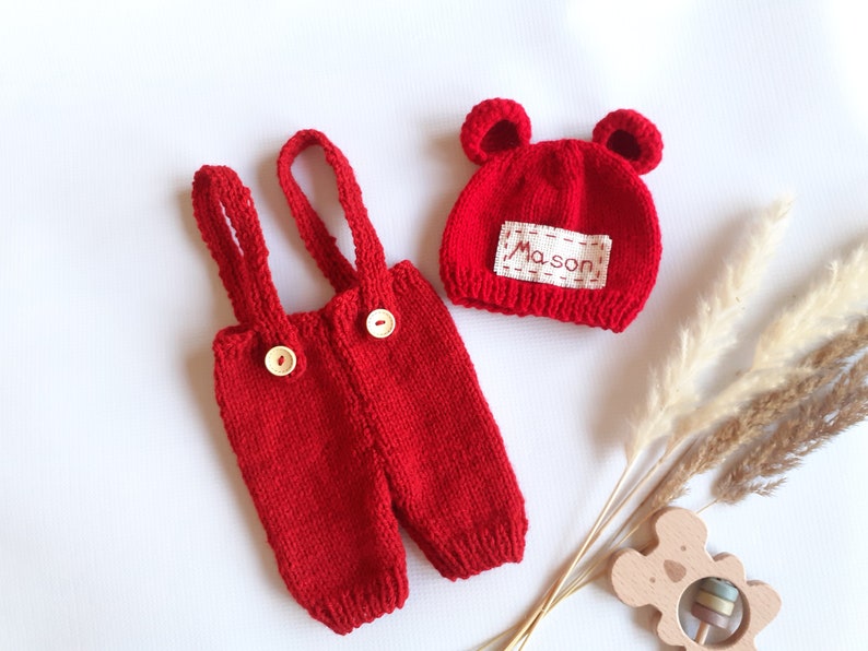 May include: A bright red knitted overall set with two light-colored buttons on the front straps and a matching red knitted hat featuring two small rounded ears and a white rectangular patch with the name Mason embroidered in red thread.