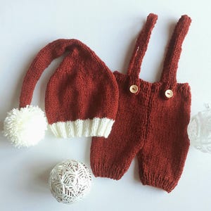 May include: A knitted rust-red outfit consisting of a long-tailed hat with a white pom-pom and a white ribbed brim, paired with matching rust-red knit overalls featuring two shoulder straps and two light-colored wooden buttons on the front.