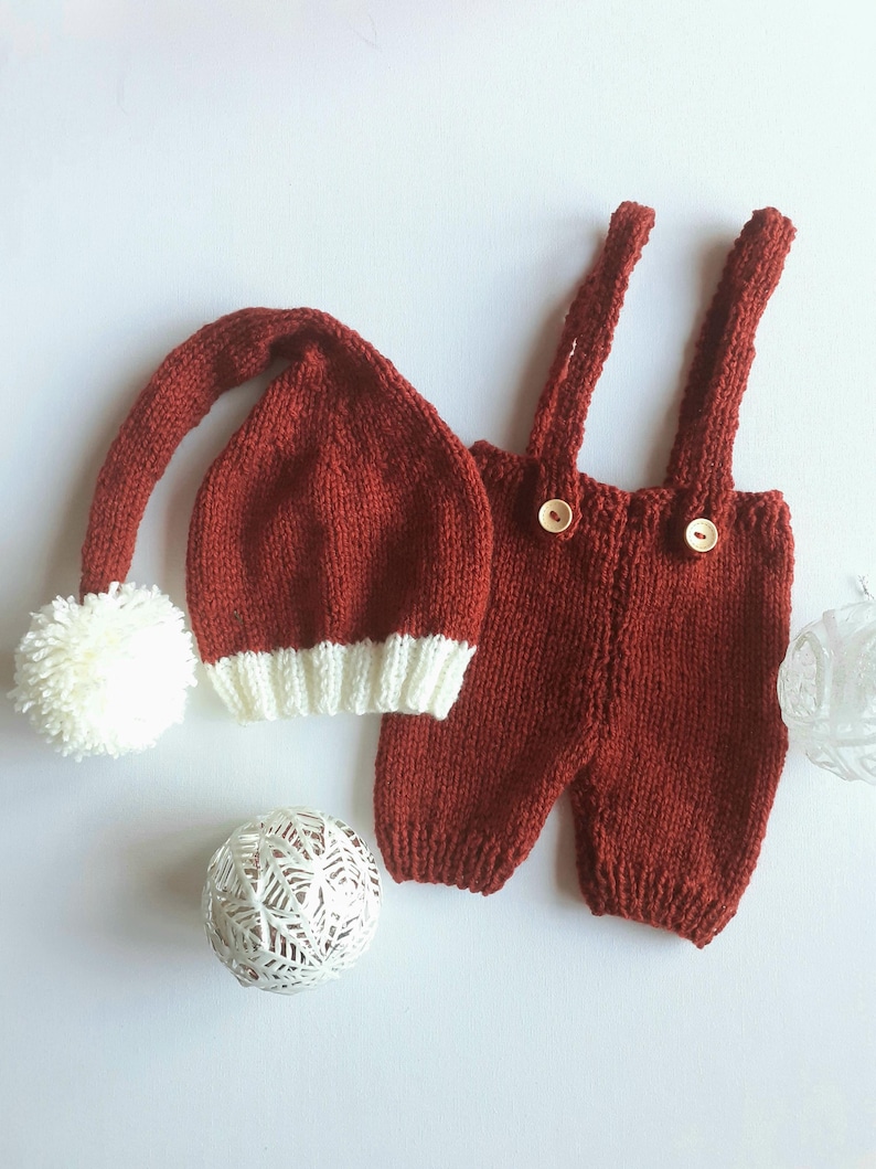 May include: A knitted rust-red outfit consisting of a long-tailed hat with a white pom-pom and a white ribbed brim, paired with matching rust-red knit overalls featuring two shoulder straps and two light-colored wooden buttons on the front.