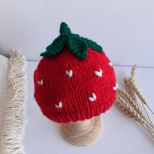 May include: A knitted red strawberry hat with green leaves and white seed details. The hat is displayed on a wooden stand. The hat is a whimsical accessory, perfect for a child or a fun-loving adult.