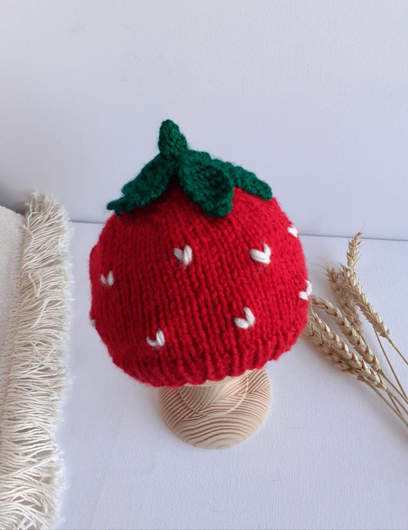 May include: A knitted red strawberry hat with green leaves and white seed details. The hat is displayed on a wooden stand. The hat is a whimsical accessory, perfect for a child or a fun-loving adult.