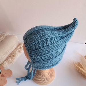 Puede incluir: Un gorro de punto azul con una parte superior puntiaguda y textura acanalada. El gorro presenta largos lazos de hilo trenzado que cuelgan de los lados, diseñados para atarse bajo la barbilla. El patrón de punto consiste en nervaduras verticales.