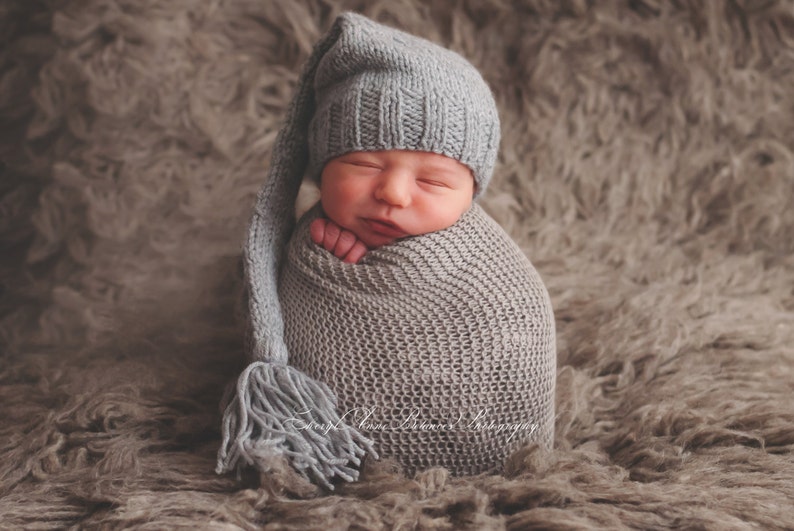 May include: A newborn baby is swaddled in a gray knitted blanket and wearing a gray knitted hat with a long tassel. The baby is sleeping peacefully.