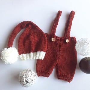 May include: A dark red knitted hat with a long tail, a white pom-pom, and a white ribbed brim, paired with matching dark red knitted overalls featuring two shoulder straps and two small wooden buttons on the front waistband.