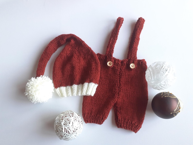 May include: A dark red knitted hat with a long tail, a white pom-pom, and a white ribbed brim, paired with matching dark red knitted overalls featuring two shoulder straps and two small wooden buttons on the front waistband.