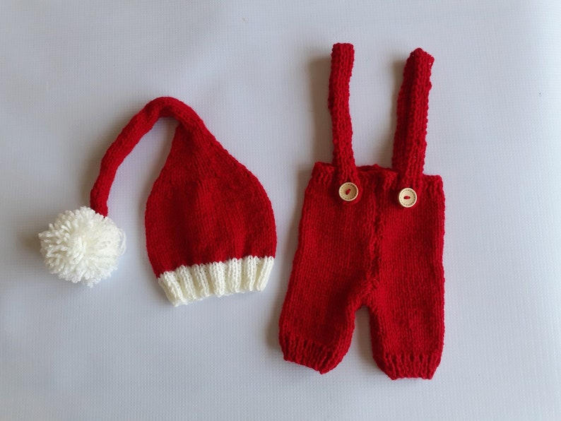 May include: A red knitted hat with a white pom-pom and a white ribbed brim, paired with red knitted overalls featuring two light-colored wooden buttons on the front straps. The set is crafted from a textured yarn in a vibrant solid red color.