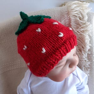 May include: A red knitted baby hat shaped like a strawberry, complete with green leaves and white seed details. The hat is worn by a baby wearing a white onesie. The hat is a fun accessory for babies.