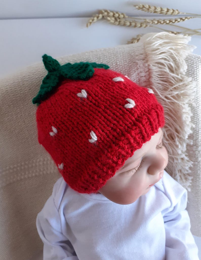 May include: A red knitted baby hat shaped like a strawberry, complete with green leaves and white seed details. The hat is worn by a baby wearing a white onesie. The hat is a fun accessory for babies.