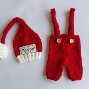 May include: A red knitted hat with a white pom-pom and a white brim featuring the name Mason, paired with red knitted overalls that have two shoulder straps and two light-colored wooden buttons on the front of the waistband.