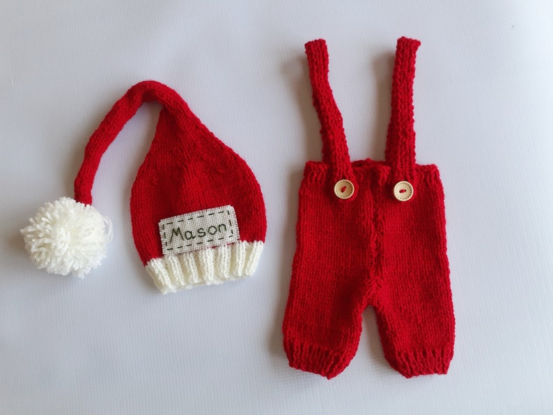 May include: A red knitted hat with a white pom-pom and a white brim featuring the name Mason, paired with red knitted overalls that have two shoulder straps and two light-colored wooden buttons on the front of the waistband.
