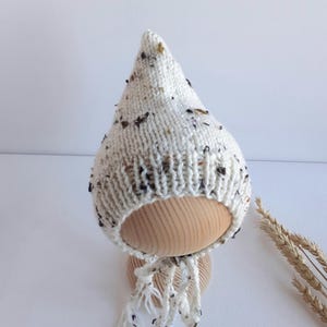 May include: A pointed knit hat in an off-white color featuring a speckled pattern with dark brown and black flecks throughout the yarn. The hat has a ribbed brim and two braided ties hanging from the bottom, crafted from the same speckled yarn.