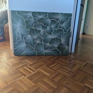 May include: A square canvas artwork with a repeating pattern of white fan-like shapes on a dark green background. The artwork is propped against a white wall and door frame, with a wooden parquet floor in the foreground.