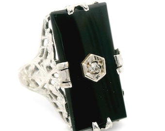 May include: A silver ring featuring a rectangular black onyx stone with a small diamond accent in a hexagonal setting. The ring has an ornate, filigree design on the band and around the onyx.
