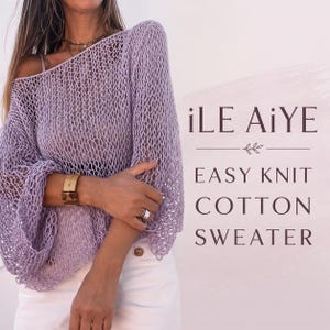 May include: A light purple, loose-knit, open-weave sweater with long sleeves and a wide neckline. The text on the right side reads: iLE AiYE, EASY KNIT COTTON SWEATER. The sweater is worn over white bottoms, featuring a visible brown button at the waist.