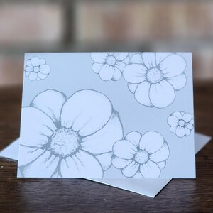 May include: A light blue greeting card featuring a floral design. The card displays several large, detailed flower illustrations in shades of gray. The card is resting on a white envelope, with a wooden surface in the foreground.