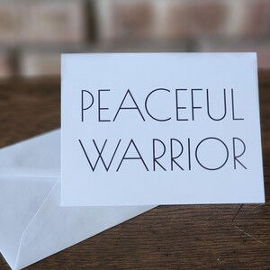 May include: A white greeting card with the words "PEACEFUL WARRIOR" in black, sans-serif font. The card is next to a white envelope, both on a wooden surface. The background includes a brick wall.