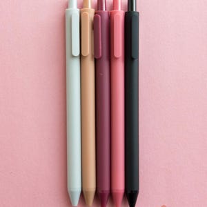 Könnte beinhalten: Fünf Stifte in verschiedenen Farben: hellgrau, beige, burgunderrot, rosa und schwarz. Die Stifte sind auf einem rosa Hintergrund aufgereiht.