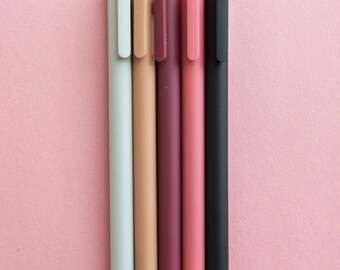 May include: Five pens in different colors: light gray, beige, burgundy, pink, and black. The pens are lined up on a pink background.