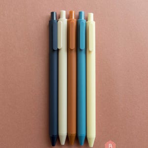 Könnte beinhalten: Fünf Stifte in verschiedenen Farben: marineblau, creme, braun, blaugrün und beige. Die Stifte sind in einer Reihe auf einer braunen Oberfläche angeordnet.
