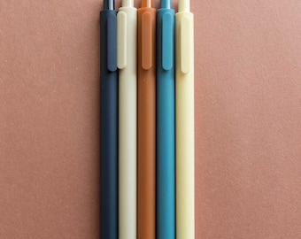 May include: Five pens in different colors: navy blue, cream, brown, teal blue, and beige. The pens are lined up in a row on a brown surface.