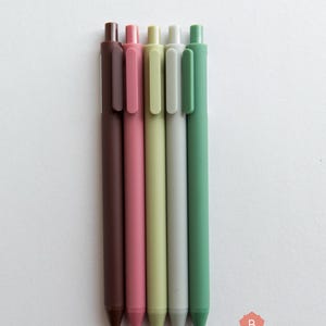 May include: Five pens in different colors: brown, pink, light green, gray, and green. The pens are all the same size and shape, and they are arranged in a row from left to right.