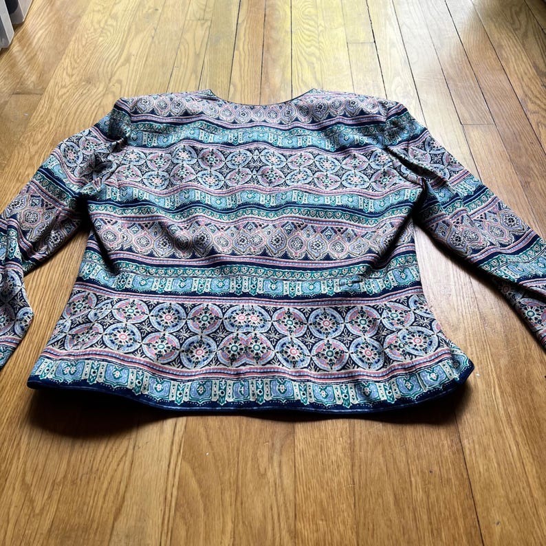Peut inclure: Veste &agrave; manches longues orn&eacute;e d'un motif color&eacute; et complexe. La veste pr&eacute;sente des motifs g&eacute;om&eacute;triques bleus, roses et blancs. Le v&ecirc;tement a un col rond et est fait d'une mati&egrave;re douce.