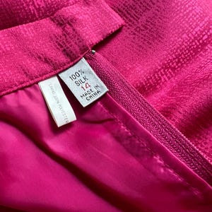 Peut inclure: Gros plan sur une jupe fuchsia vibrante avec un ext&eacute;rieur en soie textur&eacute;e et une doublure lisse en polyester rose. La jupe est dot&eacute;e d'une fermeture &eacute;clair et d'une &eacute;tiquette indiquant la taille 14, fabriqu&eacute;e en Chine, et compos&eacute;e &agrave; 100 % de soie. La doublure est en 100 % polyester.