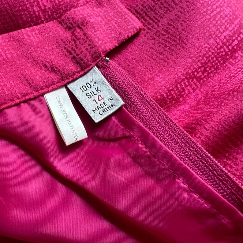 Peut inclure: Gros plan sur une jupe fuchsia vibrante avec un ext&eacute;rieur en soie textur&eacute;e et une doublure lisse en polyester rose. La jupe est dot&eacute;e d'une fermeture &eacute;clair et d'une &eacute;tiquette indiquant la taille 14, fabriqu&eacute;e en Chine, et compos&eacute;e &agrave; 100 % de soie. La doublure est en 100 % polyester.